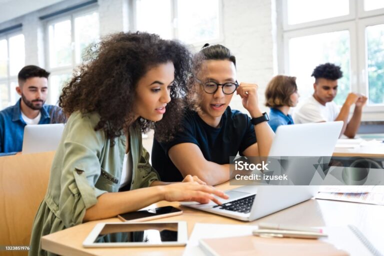 Young curly hair woman discussing with man in classroom. Classmates are learning through laptop. They are wearing casuals.
